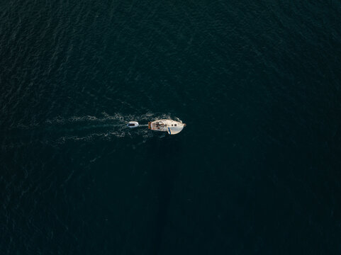 Aerial view of a white sailboat towing a small dinghy across the deep blue sea in Syracuse, Sicily, Italy.