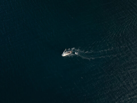 Aerial view of a white sailboat with a small dinghy in tow cruising across the deep blue sea near the Island of Ortigia Syracuse, Sicily, Italy.
