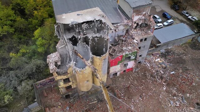 High-angle drone shot of a partially collapsed structure surrounded by forest, with an excavator arm reaching across the remains of the building and water being used to suppress dust during demolition