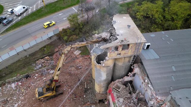High-angle drone shot of a partially collapsed structure surrounded by forest, with an excavator arm reaching across the remains of the building and water being used to suppress dust during demolition