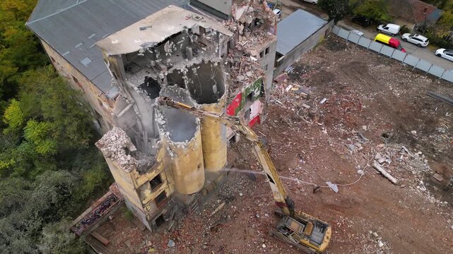 Aerial top-down view of a demolished building site with a hydraulic excavator spraying water onto rubble, broken concrete walls, twisted debris, and surrounding trees, suggesting controlled cleanup or