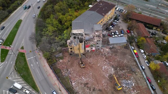 Aerial top-down view of a demolished building site with a hydraulic excavator spraying water onto rubble, broken concrete walls, twisted debris, and surrounding trees, suggesting controlled cleanup or