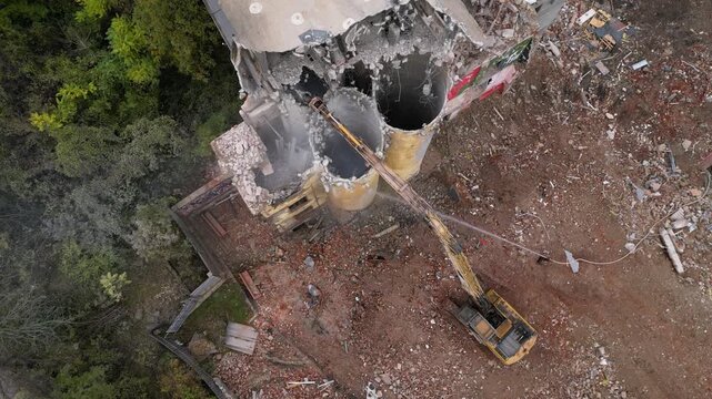 Aerial view of a partially demolished building with an excavator, rubble-strewn lot, and adjacent residential street. Destruction of property by industrial machinery.