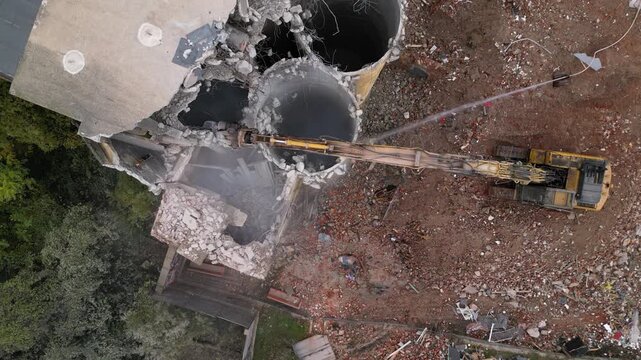 Aerial view of a partially demolished building with an excavator, rubble-strewn lot, and adjacent residential street. Destruction of property by industrial machinery.