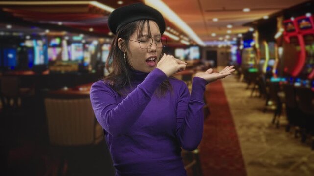 Woman pinched nose with fingers and displays open palm beside slot machines in a casino building, wearing beret and purple turtleneck; disgust playful.