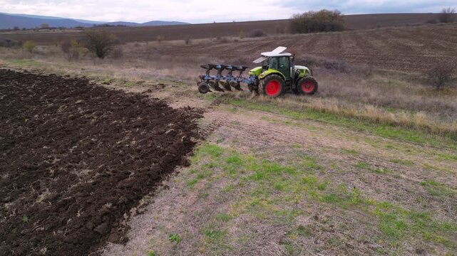 Aerial drone footage following a modern tractor pulling a mounted plow while turning soil across a large agricultural field. The clip highlights seasonal land preparation, mechanized farming, soil cul