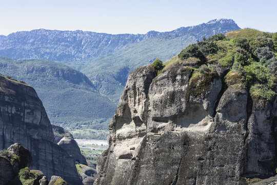 majestic sandstone cliffs of meteora, greece, under a clear blue sky, featuring dramatic rock formations, lush greenery, and the pindus mountains in the background