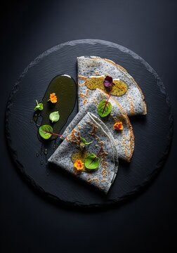 Thin gourmet crepes featuring a unique textured stone like surface, arranged on a dark slate plate with golden honey and fresh botanicals, drizzle, nature, culinary