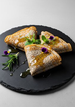 Thin gourmet crepes featuring a unique textured stone like surface, arranged on a dark slate plate with golden honey and fresh botanicals, cooked, slate, drizzle