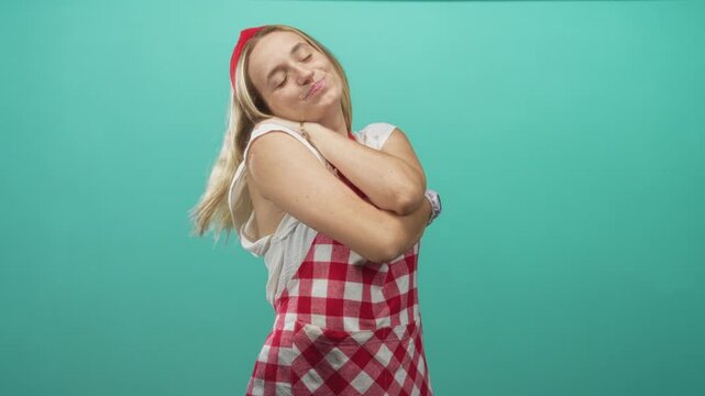Woman cook smiling and hugging her arms while wearing a red checkered apron in a studio with teal backdrop; contentment self love.