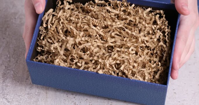 Woman opening gift box with shredded paper filler at light grey table, closeup