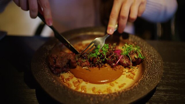 Close up of a gourmet fine dining dish being cut with fork and knife. Elegant plating with tender meat, creamy sauce, and fresh microgreens in a stylish restaurant setting