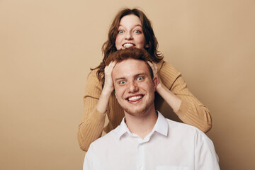 Fototapeta premium Happy young man and woman playfully pose together on beige background. The woman stands behind the man with hands on his head, both smiling joyfully. Concept of friendship and fun.