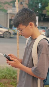 Male schoolboy in eyeglasses with backpack walking while using smartphone in city. Vertical shot of candid people, authentic mobility, modern lifestyle, communication and daily routine concept.