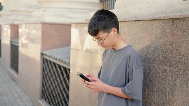 Young person standing in grey t-shirt near wall and using smartphone in relaxed urban environment. Male person order taxi via smartphone. Communication and everyday city life concept.