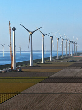 Aerial view of a row of modern wind turbines along a coastal dike with agricultural fields and a construction crane under a clear blue sky in Netherlands.