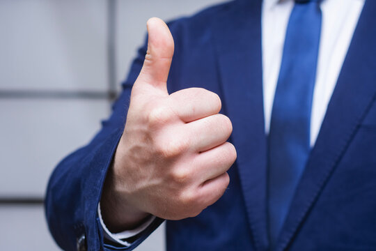 Businessman in navy suit and blue tie showing thumbs up gesture toward camera. Concept for business approval, success, positive feedback, and agreement.