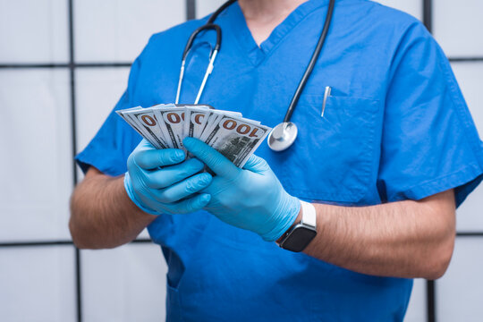 Doctor in blue scrubs and latex gloves holding fan of 100 dollar bills with stethoscope. Concept for healthcare cost, medical fee and hospital payment.