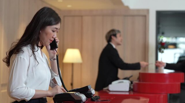A professional female receptionist speaks on a landline phone to confirm guest reservations at a luxury hotel front desk counter.
