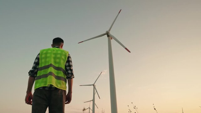 Engineer in safety vest standing near wind turbine farm at sunset looking at alternative energy generation.