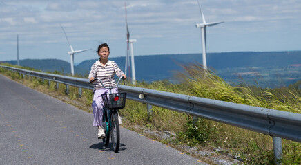 Obraz premium Smiling Asian girl riding bicycle along rural road with wind turbines and mountain view, concept of eco-friendly lifestyle.