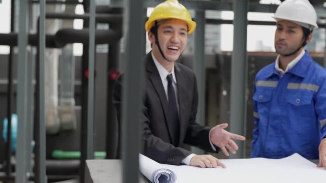 An Asian construction consultant in a suit and a technician in hardhats review MEP piping blue-prints and schematic drawings on an outdoor building site.