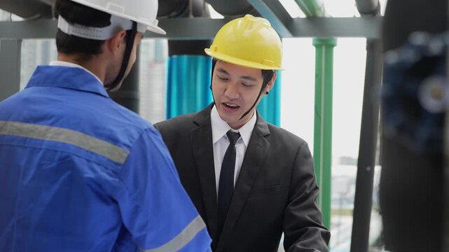 An Asian construction consultant in a suit and a technician in hardhats review MEP piping blue-prints and schematic drawings on an outdoor building site.