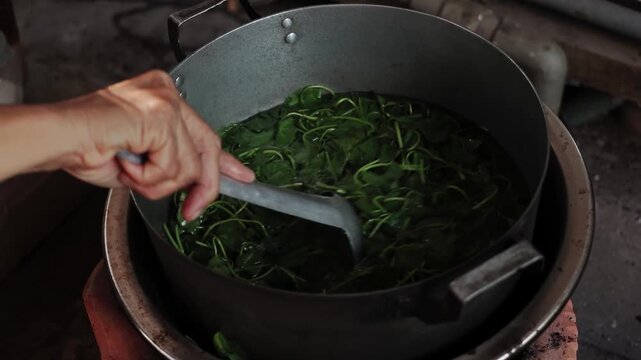 A woman stirring fresh pennywort leaves in a boiling pot on a traditional stove in a rustic Vietnamese kitchen