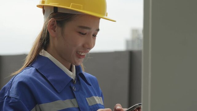 A female electrical engineer wearing a yellow hardhat and blue uniform operates a digital control panel display while holding a smartphone during a technical building system audit.