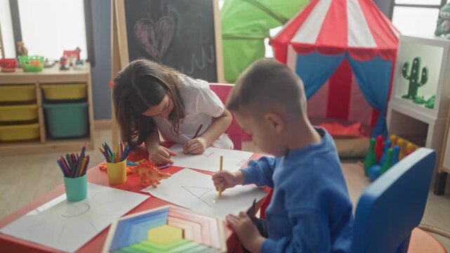 Girl leans over paper and boy holds pencil drawing with colored pencils at studio table with toy tent and chalkboard; calm concentration.