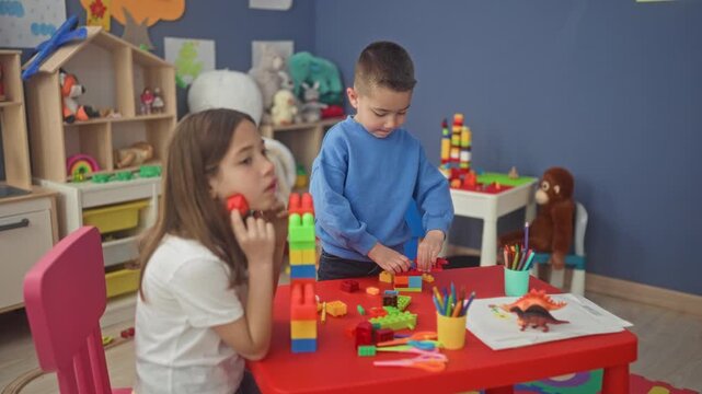Boy stacking colorful plastic blocks with hands on a red table in a playroom inside a building while girl watches and crayons sit in a cup; quiet concentration.