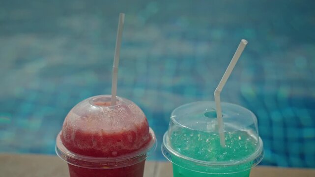 Two refreshing frozen drinks with straws, one red and one blue, sit by edge of a sparkling blue swimming pool on a sunny day.