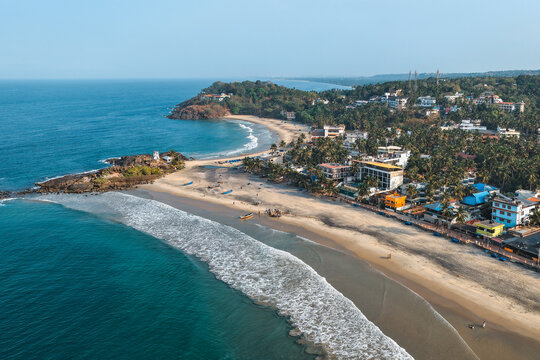 Aerial view of the crescent-shaped sandy coastline with turquoise waves, palm-fringed hotels, and rocky headlands under a clear blue sky in Kovalam, Kerala, India.