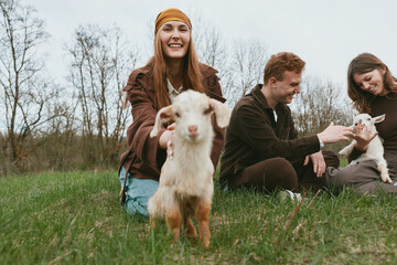 Fototapeta premium A cheerful group enjoys a sunny day on a field, with a playful goat kid approaching the camera. A candid lifestyle moment showing bonding, exploration, and gentle animal interaction.