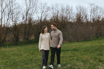 Fototapeta premium Young couple wearing knitted sweaters walks together on a green meadow in early spring with bare trees in the background, enjoying a calm and intimate moment outdoors.