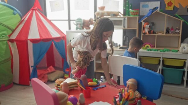 Girl leans over doll and arranges toy food on a red table while a boy sorts toys from storage bins in a bright children's playroom inside a building; sibling playfulness.