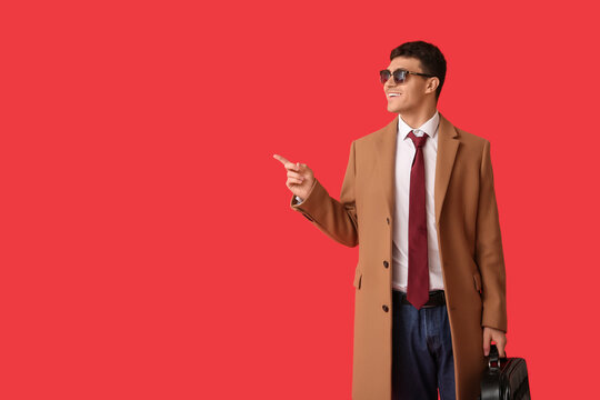 Smiling young man in coat and stylish tie with sunglasses pointing at something on red background