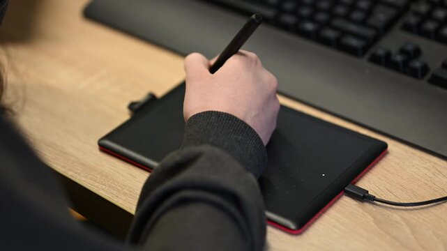 Close up of a student's hand using a black stylus on a compact pen tablet connected by USB cable