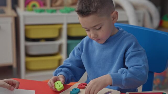 Boy placing wooden number puzzle pieces with hands at preschool classroom table; concentration learning.