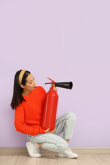 Young Asian woman with fire extinguisher sitting against lilac wall