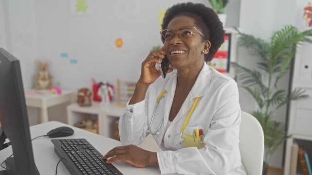 Woman doctor in coat holding phone to ear while typing at a computer on a clinic desk in a building; compassionate care support.