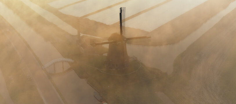 Aerial view of De Kaagmolen windmill surrounded by misty polder fields and canals at sunrise in Spanbroek, North Holland, Netherlands.