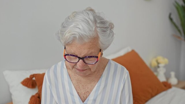 Elderly woman wearing colorful glasses sits on bed and looks at camera among orange pillows and a vase on a bedside table in building; calm reflection.