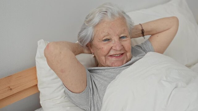 Woman senior lying in bed with hands behind head, smiling, pearl earring and bracelet, pillow and duvet visible in building; serenity comfort morning wakeup.