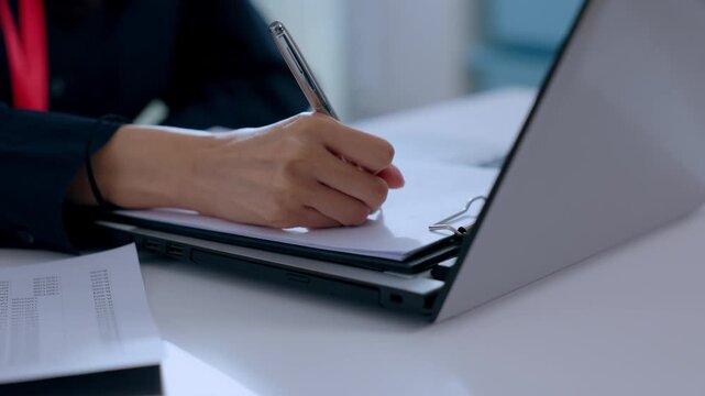 Focused business person writing document with pen in hand at modern office desk next to closed laptop showing professional concentration during corporate paperwork task