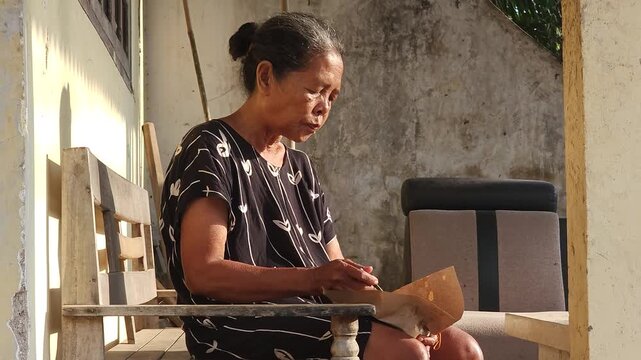 An elderly Asian woman sits on a wooden bench outdoors, eating from a paper wrapper and talking in the sunlight.