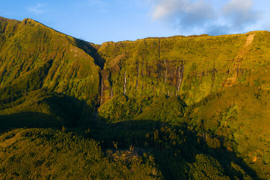 Aerial view of Poco da Ribeira do Ferreiro waterfalls cascading down lush green cliffs into a dense forest during golden hour Fajazinha, Azores, Portugal.