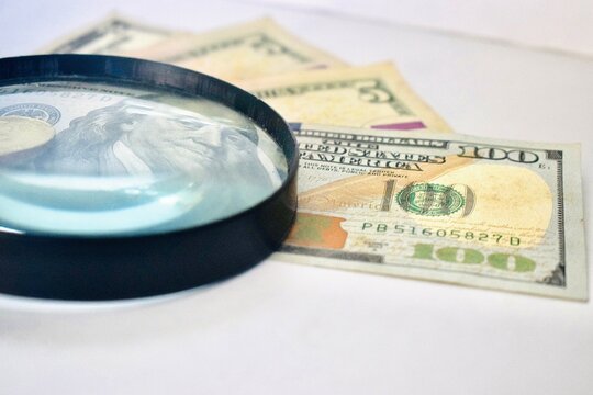Close-up of a magnifying glass highlighting the detail of a 100 US dollar bill, representing financial scrutiny, counterfeiting detection, or economic analysis.