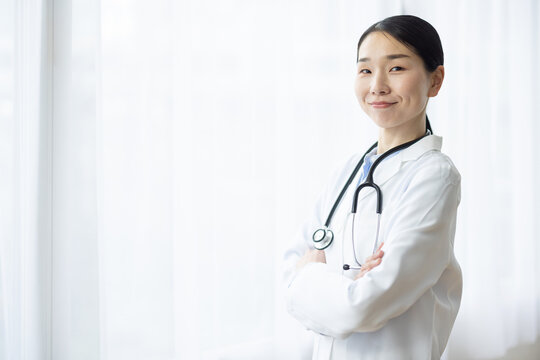 Asian Female Doctor in White Coat Smiling with Arms Crossed by Bright Window