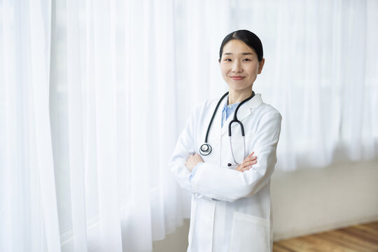 Asian Female Doctor in White Coat Smiling with Arms Crossed by Bright Window